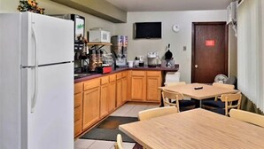 A compact kitchen with wooden cabinets, a white refrigerator, a microwave, and a coffee maker. There is a dining table with chairs and a TV mounted on the wall.