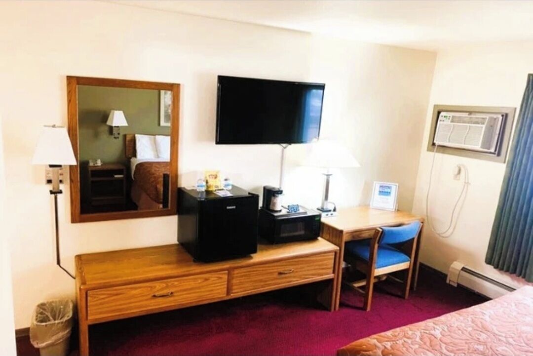 Una habitación de hotel con una cama, un escritorio con silla, televisor, espejo, lámpara, calentador y un cubo de basura.