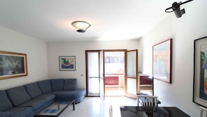 Una sala de estar con un sofá azul, una mesa de centro y una mesa de comedor. Hay cuadros enmarcados en las paredes y una puerta corrediza de vidrio que da a un área exterior.