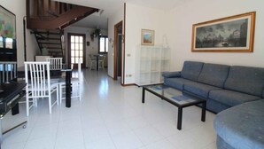 Amplia sala de estar con un sofá azul, una mesa de centro de vidrio y una zona de comedor blanca con sillas. Se ve una escalera al fondo.