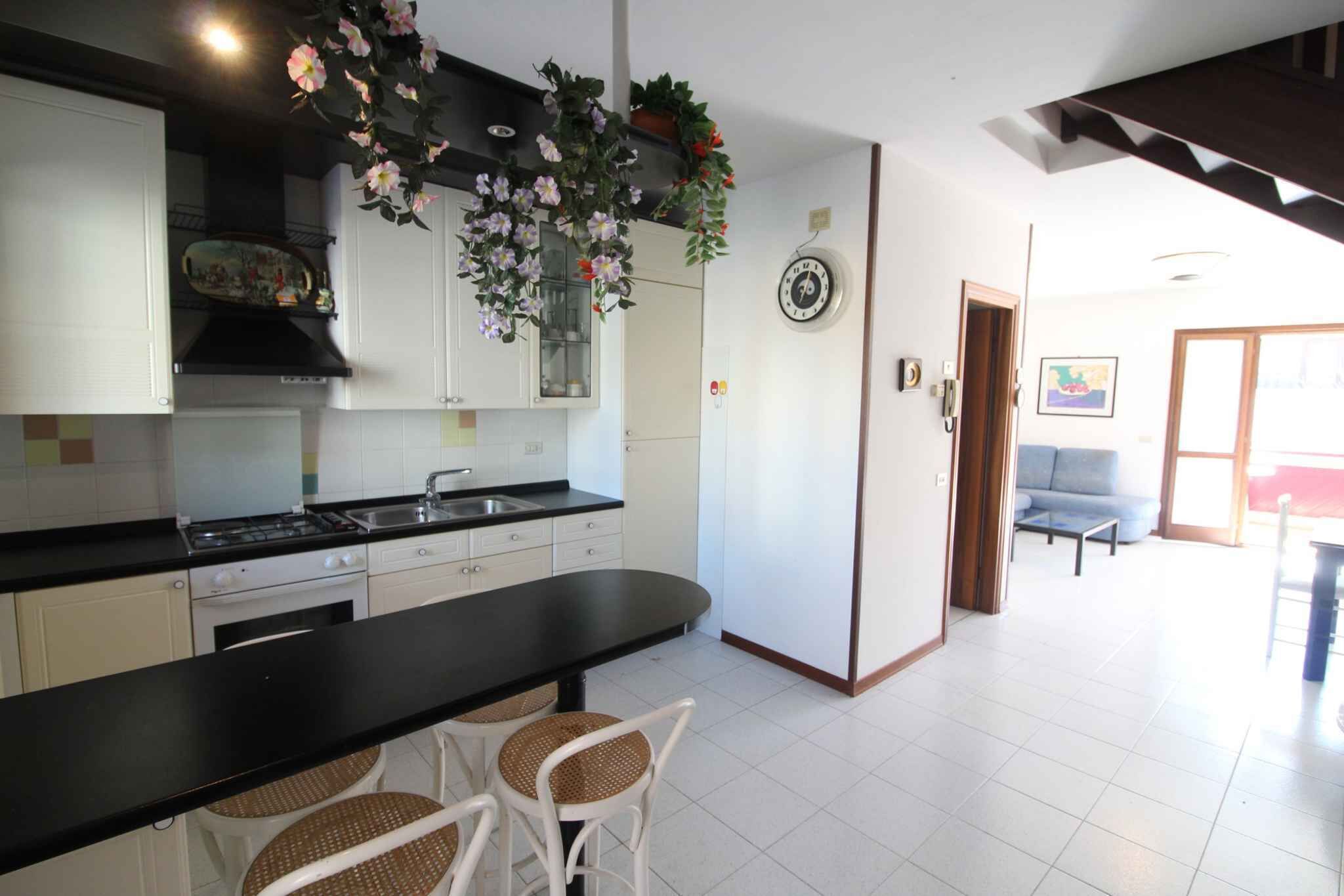 Una cocina con armarios blancos, encimera negra y una zona de comedor con sillas de mimbre.