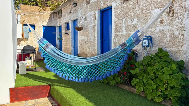 Een hangmat gespannen tussen twee gebouwen met blauwe deuren, een kleine tuin met kunstgras, en een rode bank.