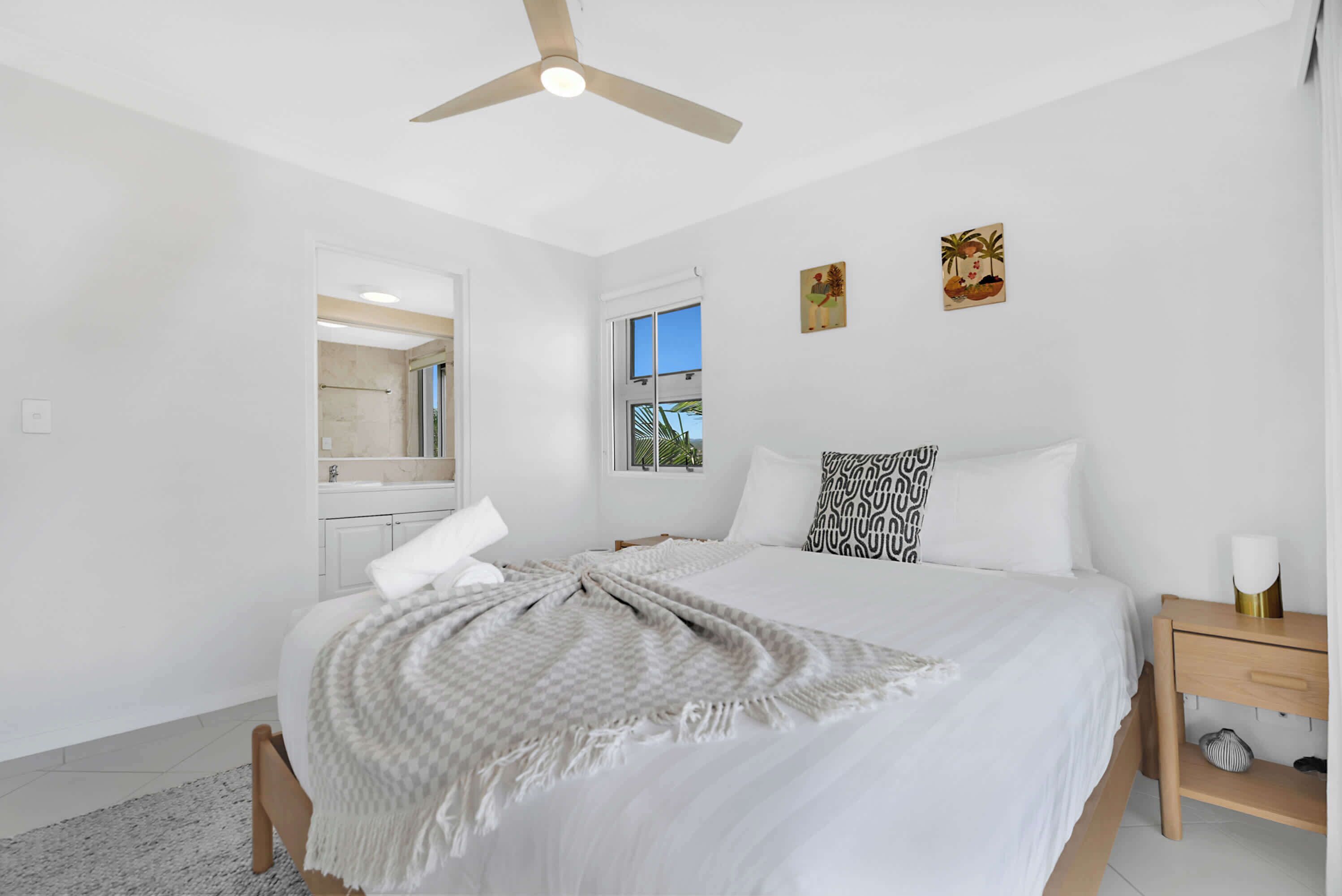 A bedroom with a bed, a nightstand, a ceiling fan, a window, and artwork on the wall.