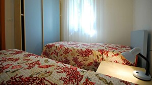 Een kamer met twee bedden, een bureau met lamp, een raam met dunne gordijnen en een deur.