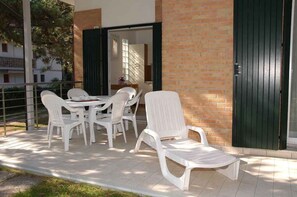 An outdoor patio with white plastic chairs and a table, a brick wall, and a green door. - Holiday apartment mit Terrasse (Lignano Sabbiadoro)
