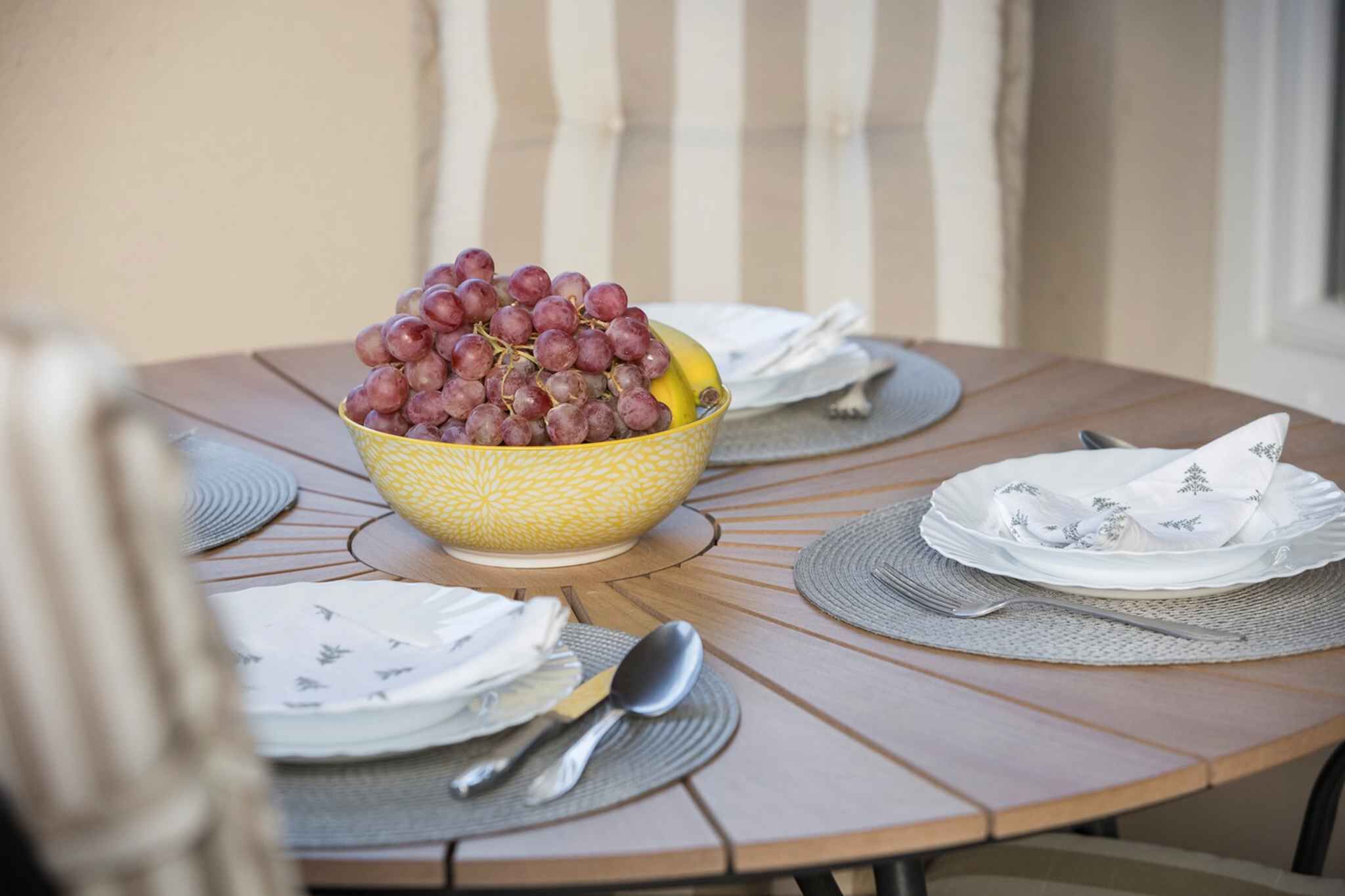 Une table ronde en bois, sur laquelle se trouve un saladier jaune rempli de raisins rouges et une banane, le tout entouré d’assiettes blanches et de couverts en argent.