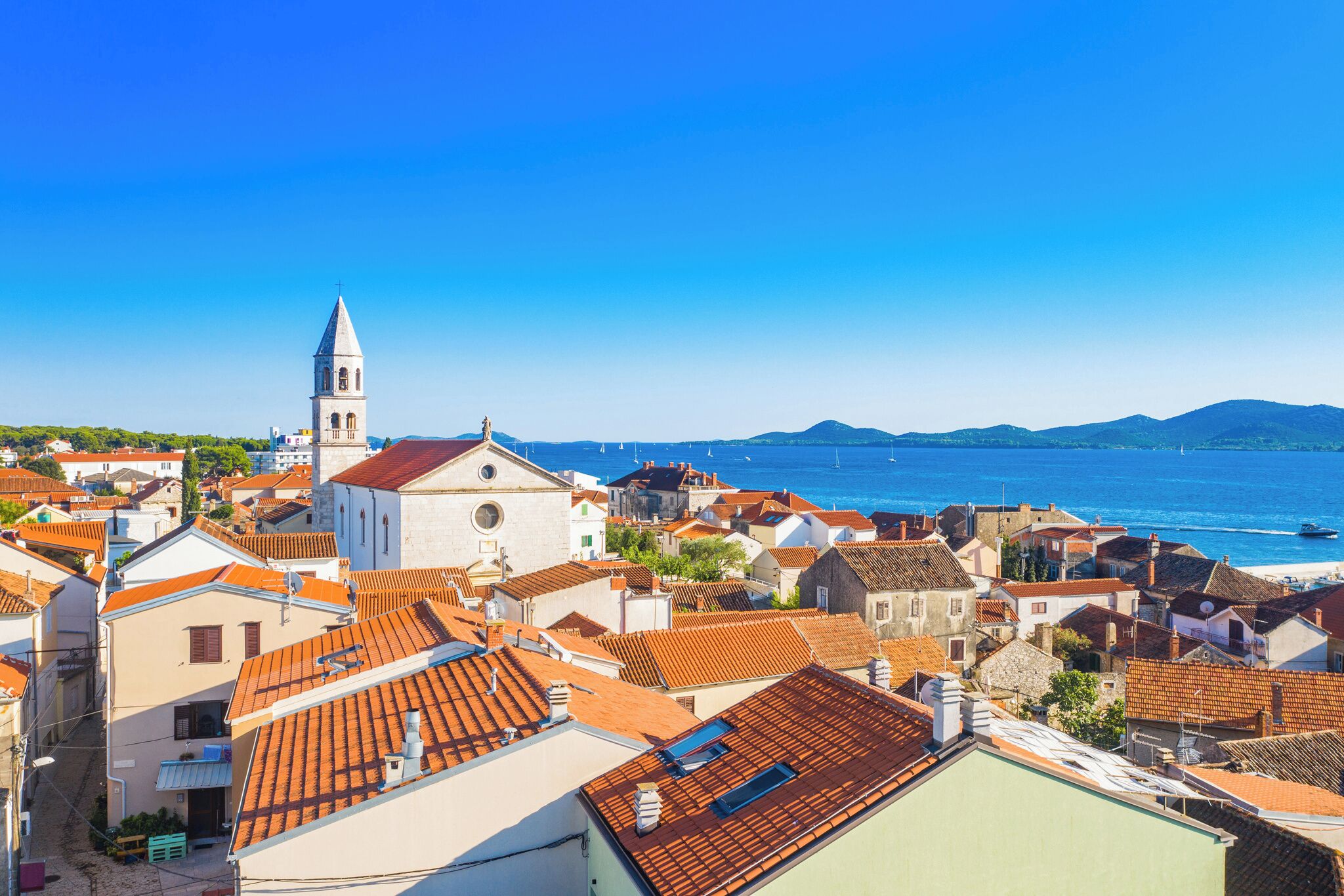 Une ville côtière aux bâtiments aux toits de tuiles rouges, dotée d’un clocher d’église imposant et offrant une vue dégagée sur la mer.