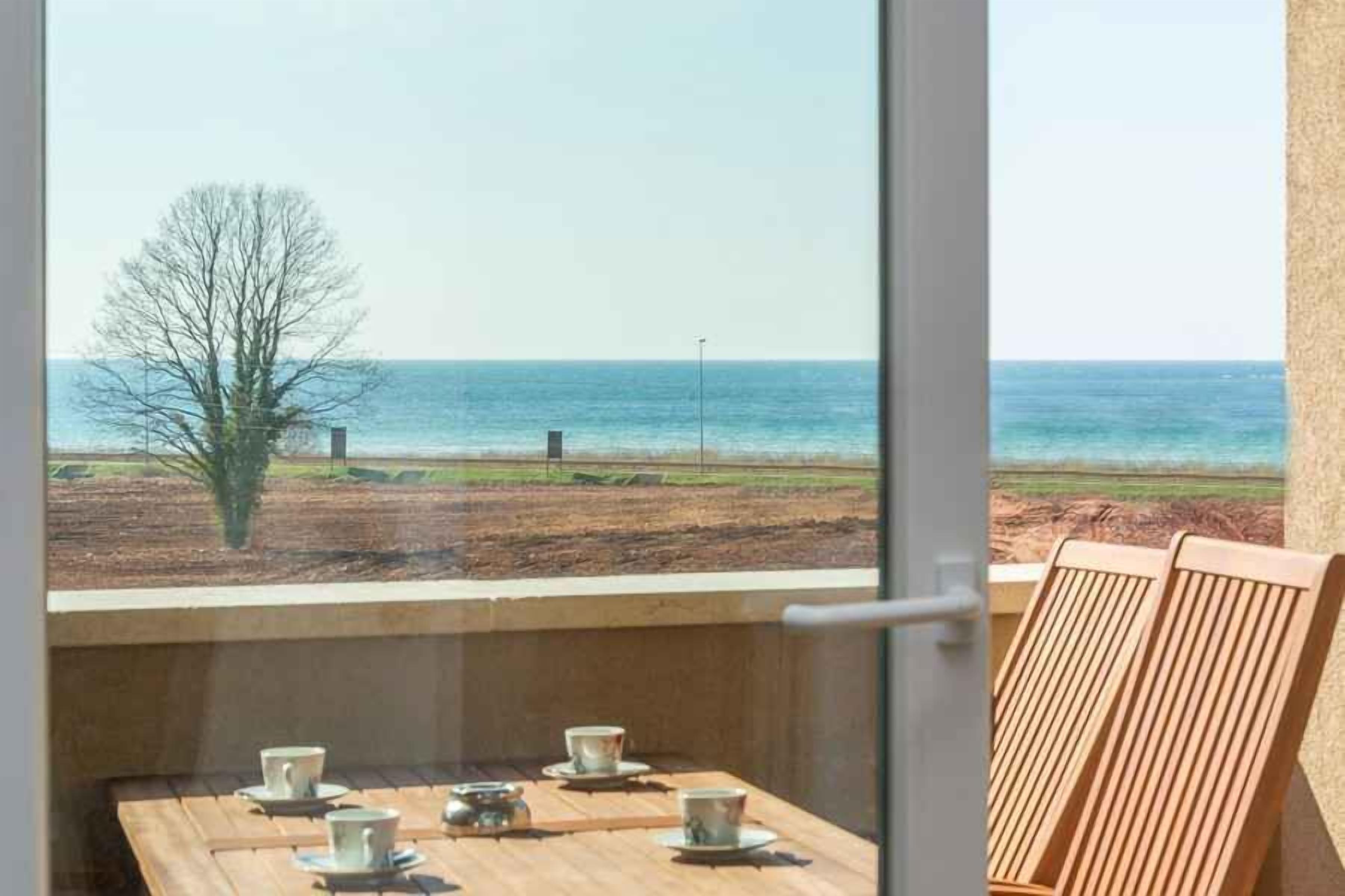A balcony with a view of the sea, a table with cups and saucers, and wooden chairs.