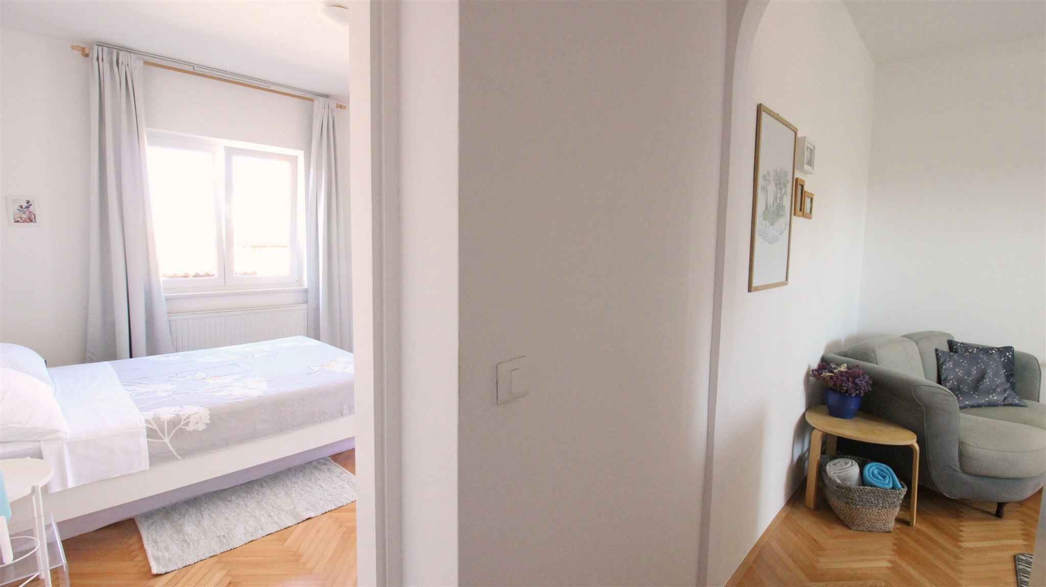 A bedroom with a bed, a window with curtains, a small table with a plant, and a grey sofa.