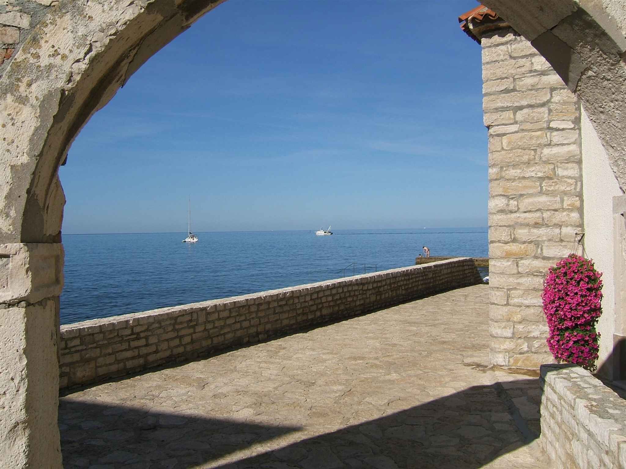 Ein steiner Torbogen mit Blick auf ein ruhiges Meer, Boote, eine Steinmauer und ein Blumenbeet.