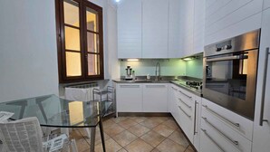 Ett kompakt kök med vita skåp, ett glasbord, ett fönster med persienner och en inbyggd ugn.