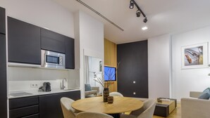 Una cocina moderna con armarios oscuros, microondas y cafetera. Hay una zona de comedor con una mesa redonda y cuatro sillas. Un televisor está montado en la pared, y se ve un sofá al fondo.