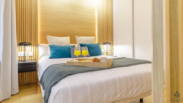 Habitación de hotel con cabecera de madera, cama con ropa de cama blanca y azul, dos lámparas de noche y una bandeja con alimentos para el desayuno.