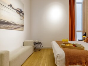 Habitación de hotel con cama, una mesita, una silla, un cuadro y ventana con cortinas.