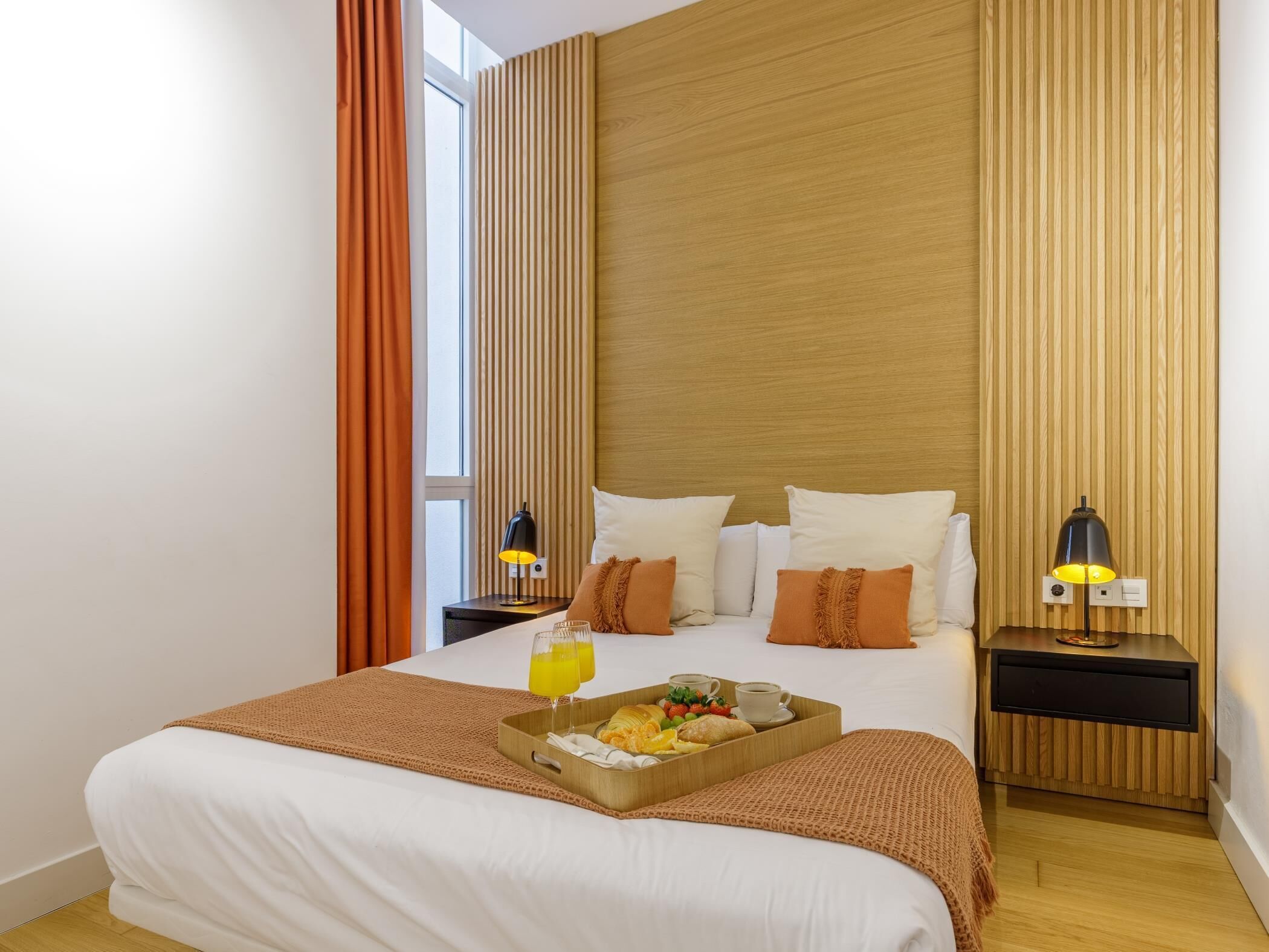 Habitación de hotel con una cama grande, dos mesitas de noche, un cabecero de madera, una ventana con cortinas y una bandeja con alimentos para el desayuno.