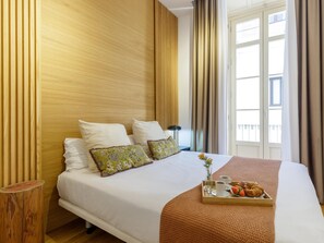 Una habitación de hotel con una cama, almohadas, una bandeja con comida y bebidas, y una ventana con cortinas.