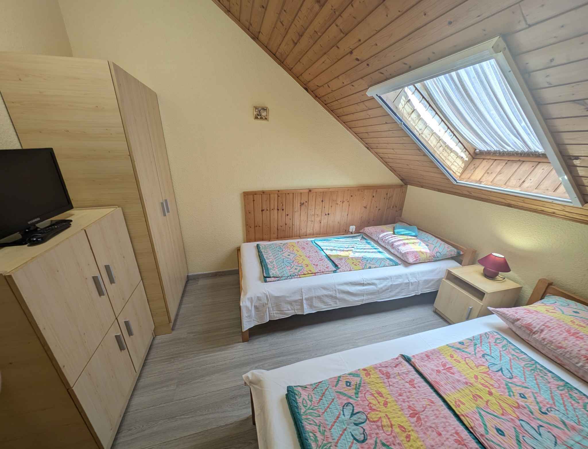 A small, cozy bedroom with a sloped ceiling, a single bed with colorful bedding, a nightstand with a lamp, and a television on a wooden cabinet.