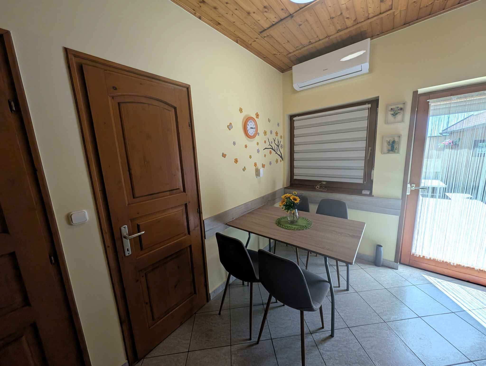 A dining area with a wooden table, two black chairs, a vase with flowers, and a wall clock.
