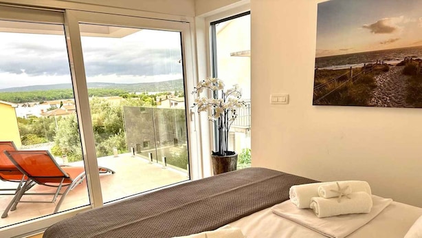 Ein Schlafzimmer mit einem großen Fenster, einem Bett mit weißer Bettwäsche, Meerblick und einem Stuhl davor.