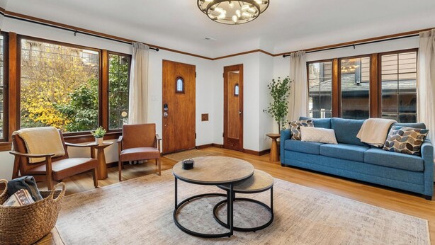 Een ruime woonkamer met een blauwe bank, houten stoelen, een ronde salontafel en grote ramen met uitzicht op bomen.
