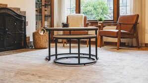 Een knus vertrek met een ronde tafel, twee stoelen, een rieten mand en een kast met glazen deuren.