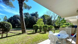 A patio with a table and chairs, surrounded by a fenced garden with trees and grass. - Holiday apartment with garden near the sea (Bibione)