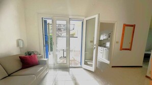 Een woonkamer met een bank, een deur naar een balkon en een zichtbare keuken.