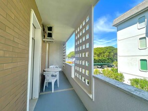 A balcony with a table and chairs, a small air conditioning unit, and a view of a parking area and greenery. - Holiday apartment near the sea (Bibione)