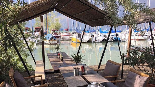 Área de refeições ao ar livre com móveis de vime e uma mesa, com vista para uma marina com barcos.