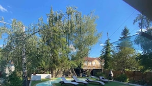 Piscina com espreguiçadeiras, cercada por árvores e um edifício ao fundo.