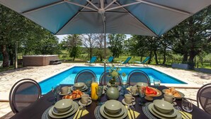 Ein Esstisch mit gedecktem Frühstück und Blick auf einen Pool, mit Gartenstühlen und einem großen Sonnenschirm.