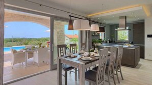 Et moderne køkken med en central ø, et spiseområde med en glasdør til en terrasse, og udsigt til en swimmingpool.