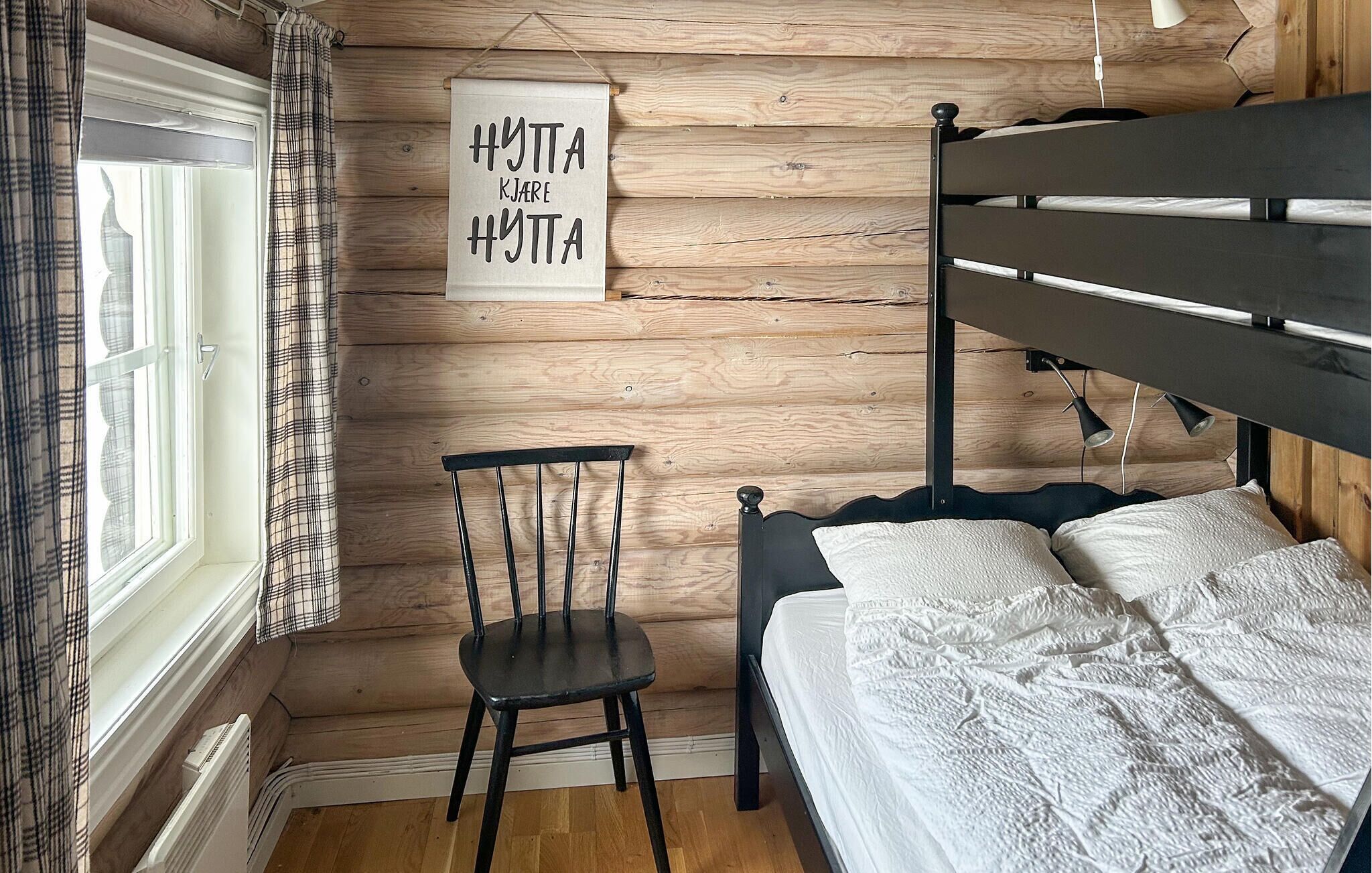 A bedroom with a wooden wall, a black metal bed frame, a white bedspread, a black chair, and a window with curtains.