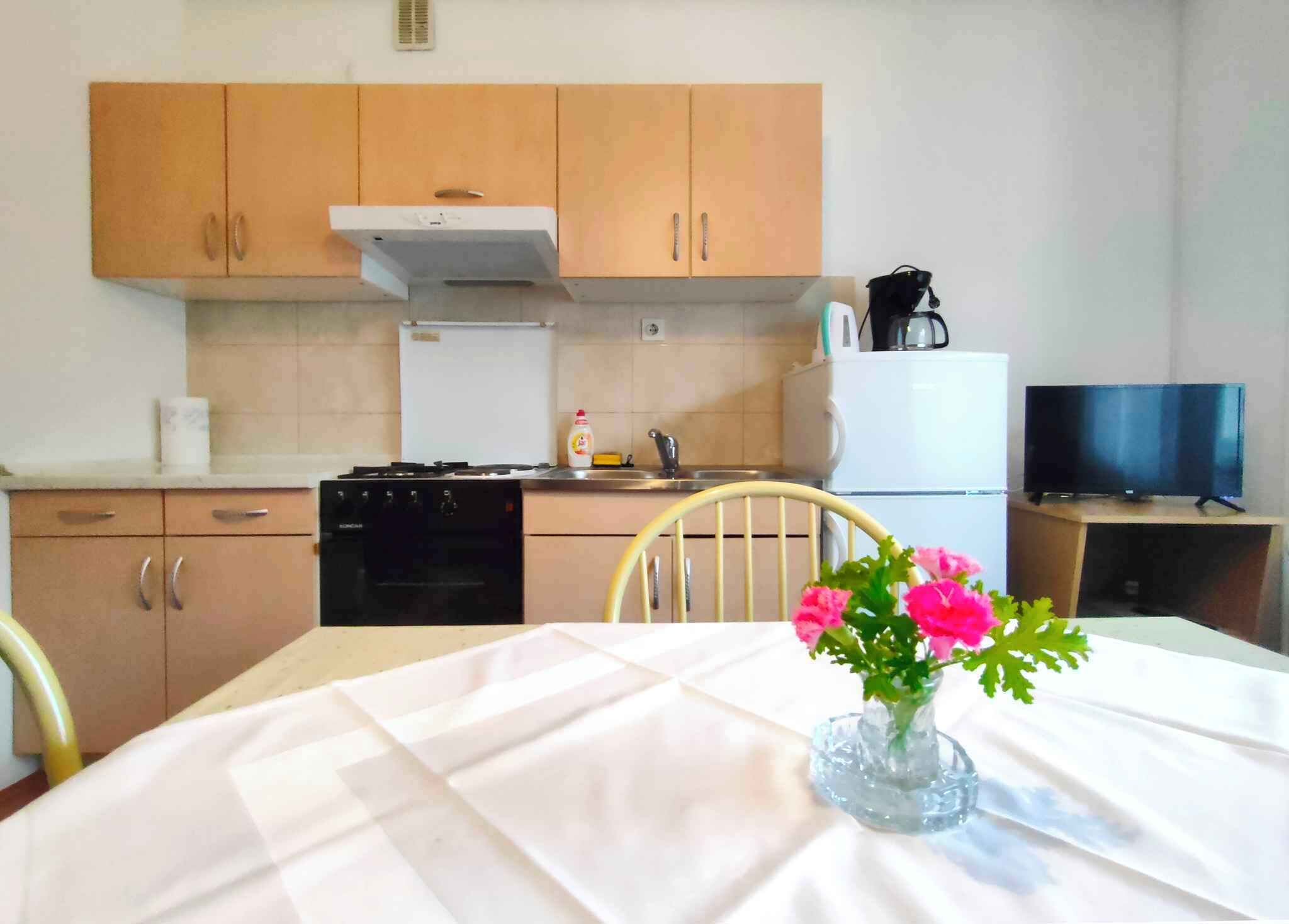Una pequeña cocina con armarios de madera, una estufa, un lavamanos y un refrigerador. Hay una mesa con un florero y una silla.