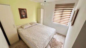 Een kleine, verzorgde slaapkamer met een bed, een kledingkast en een raam met zonwering.
