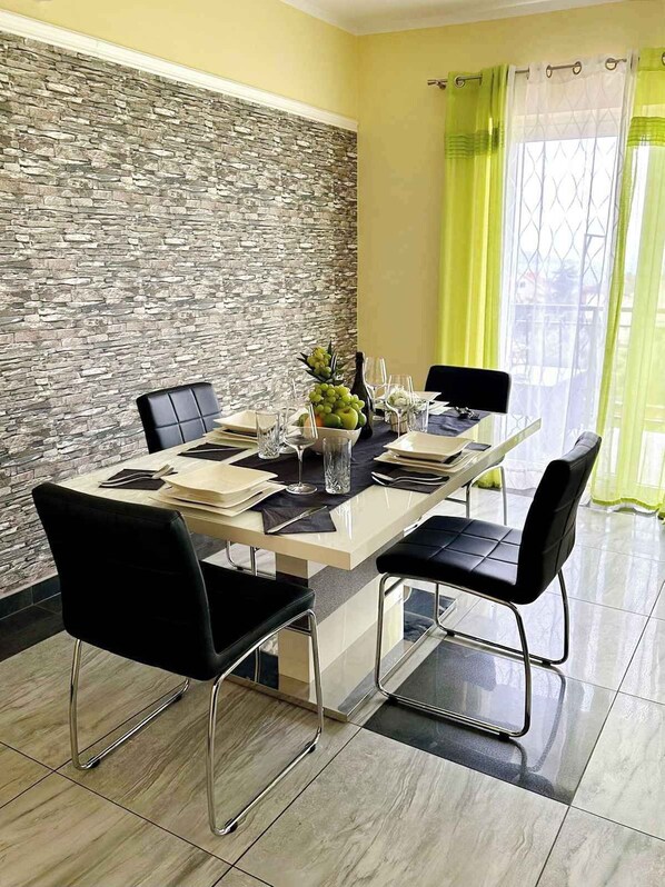 Een eetgedeelte met een stenen muur, een lange tafel gedekt voor vier personen, zwarte stoelen en uitzicht door een raam met groene gordijnen.