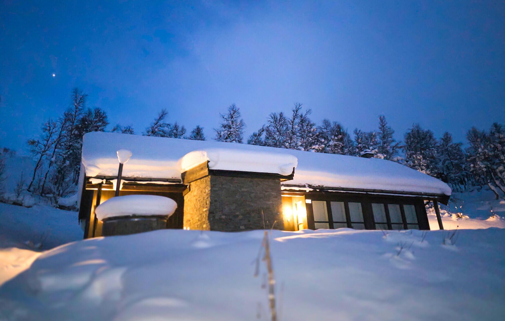 Edifici cobert de neu amb façana de pedra i porta de fusta, envoltat d'arbres carregats de neu.