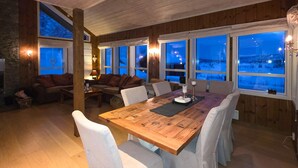 En ombonad inredning med ett matbord i trä, vita stoppade stolar, en soffa och stora fönster med utsikt över ett snötäckt landskap.