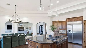 A spacious kitchen with a central island, wooden cabinets, stainless steel appliances, and a dining area with green upholstered chairs. - Epic Views | Pool • Spa • RV Garage | Near Marina (Lake Havasu City)
