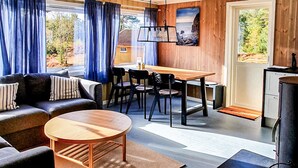 Una sala acogedora con un sofá, una mesa redonda de centro y una mesa de comedor con sillas. Hay un cuadro en la pared y vistas a zonas verdes al exterior.
