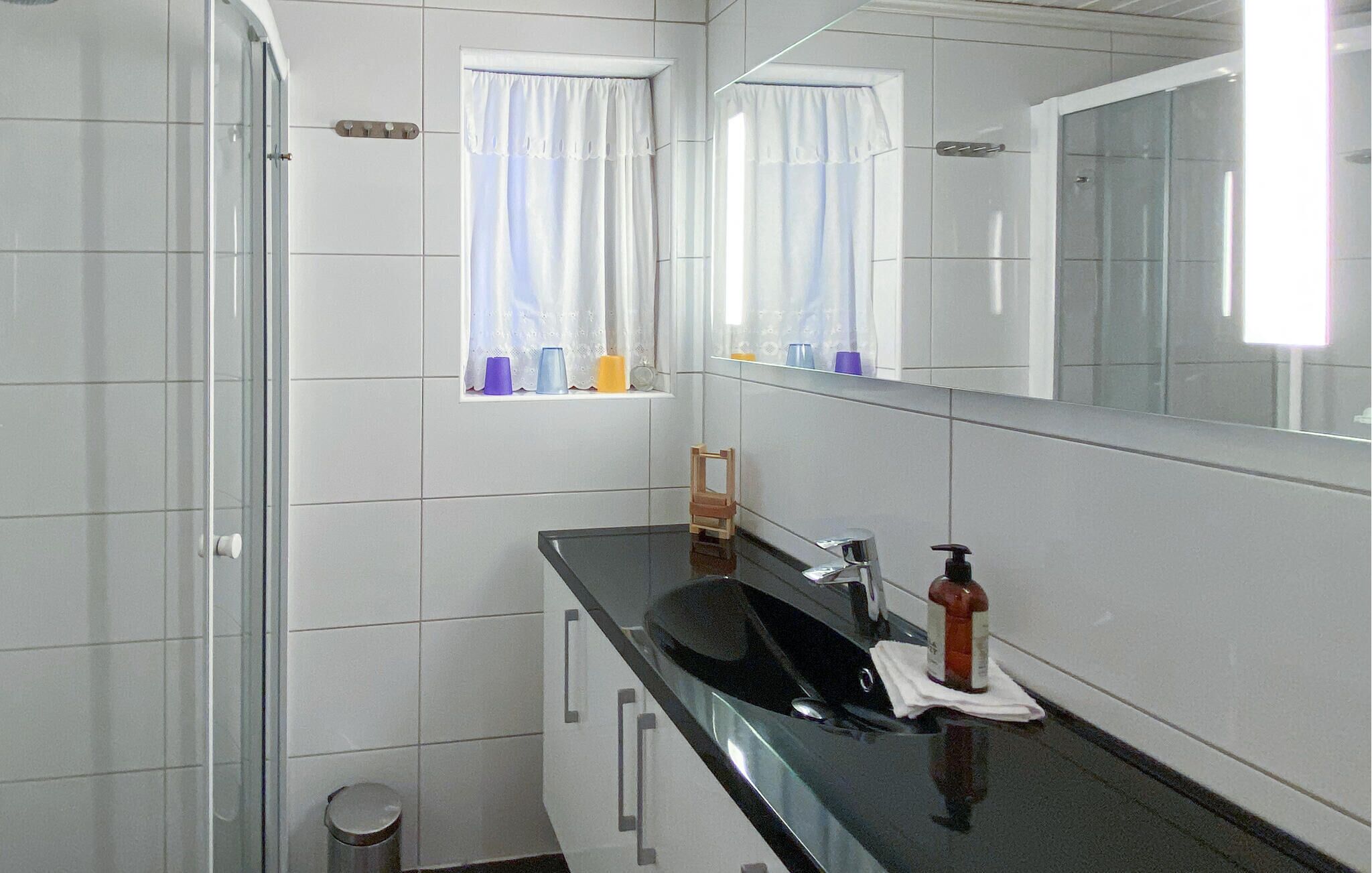 Une salle de bain avec des murs recouverts de carreaux blancs, une douche en verre, un lavabo noir avec un distributeur de savon et une fenêtre avec des rideaux.