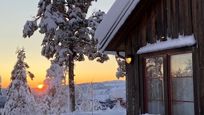 Une cabane en bois, dont le toit et les fenêtres sont recouverts de neige, entourée d’arbres chargés de neige, avec un coucher de soleil en arrière-plan.