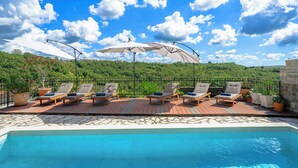 Un espace aménagé au bord de la piscine, avec des chaises longues et des parasols, entouré d’une terrasse en bois et bordé de pierres.