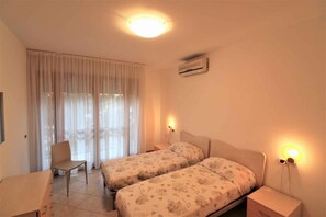 A bedroom with two beds, a chair, a desk, and a window with curtains. - Holiday apartment with air conditioning and balcony (Lignano Sabbiadoro)
