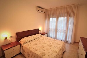 A bedroom with a bed, nightstands, an air conditioner, and a window with curtains. - Holiday apartment with air conditioning and balcony (Lignano Sabbiadoro)