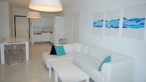 Een moderne woonkamer, ingericht in witte tinten, met een bank, een eettafel en een keuken op de achtergrond.