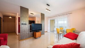 Een moderne woonkamer met een flatscreen televisie, een bank met rode kussens, een eethoek met gele stoelen, en een schuifdeur naar een balkon.