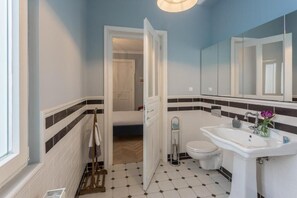 Een badkamer met een wit wastafel, een spiegel en een toilet. Er is een handdoekhouder en een deur naar een andere kamer.