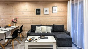 Una sala de estar moderna con una pared de madera, un sofá negro, una mesa de centro blanca y un comedor con una mesa lista para dos comensales.