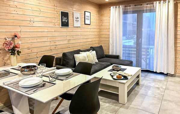 A cozy room with a wooden wall, a sofa, a dining table set with plates and glasses, and a window with curtains.